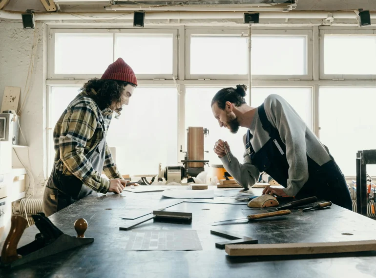 Two craftsmen collaborating on a project in a well-lit workshop, surrounded by woodworking tools and materials.