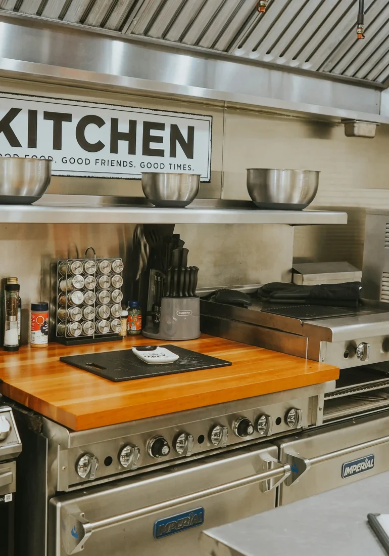 Modern kitchen with stainless steel appliances, wooden countertop, and a "KITCHEN" sign, featuring cooking utensils and equipment.