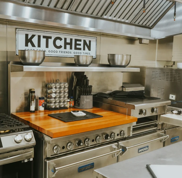 Modern kitchen with stainless steel appliances, wooden countertop, and a "KITCHEN" sign, featuring cooking utensils and equipment.