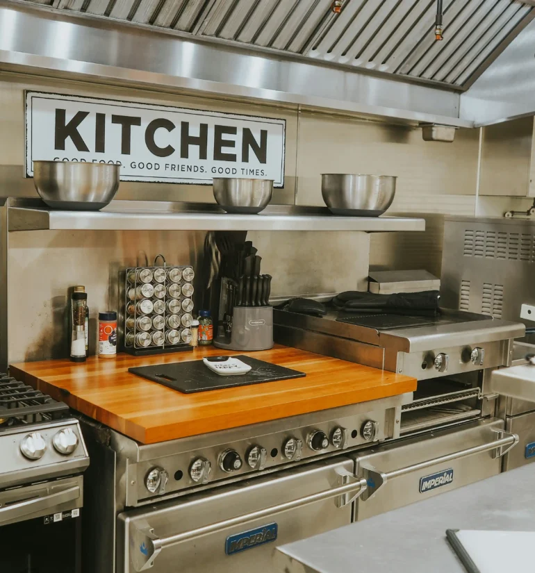 Modern kitchen with stainless steel appliances, wooden countertop, and a "KITCHEN" sign, featuring cooking utensils and equipment.