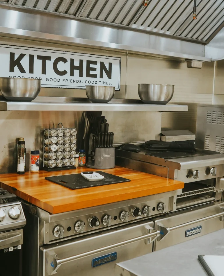 Modern kitchen with stainless steel appliances, wooden countertop, and a "KITCHEN" sign, featuring cooking utensils and equipment.