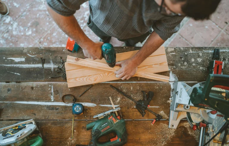 A craftsman using a drill on wooden planks in a workshop with tools and measuring equipment on a workbench.