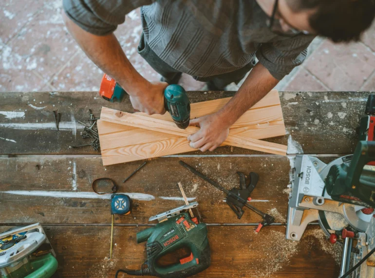 A craftsman using a drill on wooden planks in a workshop with tools and measuring equipment on a workbench.