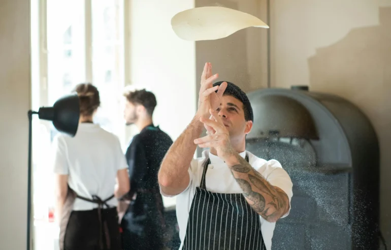 A chef tossing pizza dough in the air in a modern kitchen with a pizza oven in the background.