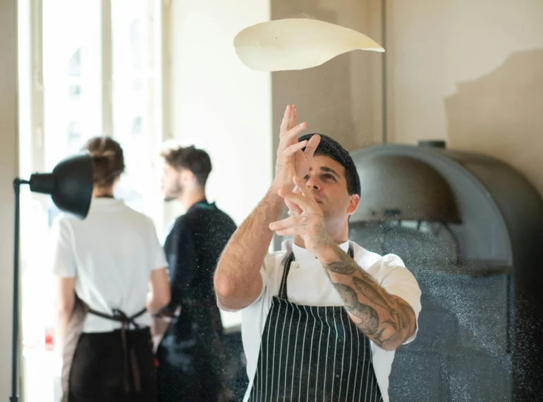 A chef tossing pizza dough in the air in a modern kitchen with a pizza oven in the background.