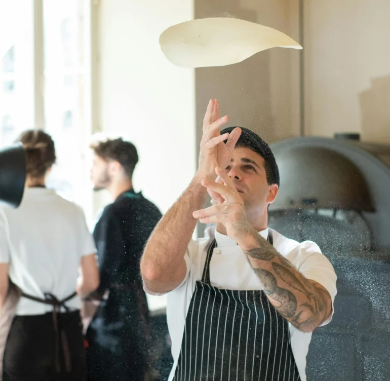 A chef tossing pizza dough in the air in a modern kitchen with a pizza oven in the background.