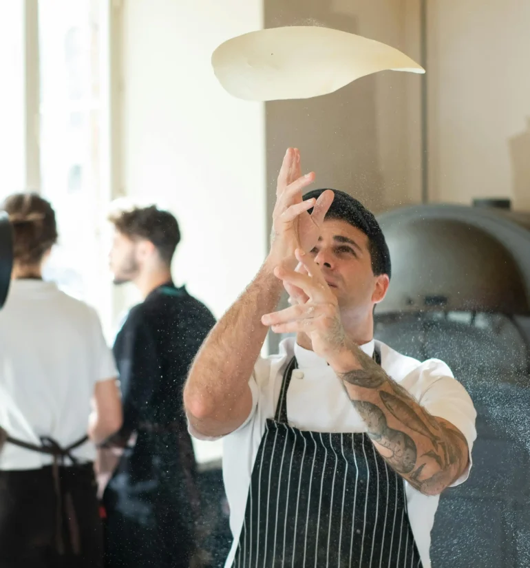 A chef tossing pizza dough in the air in a modern kitchen with a pizza oven in the background.