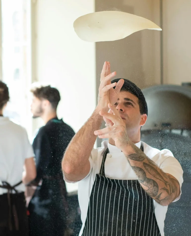 A chef tossing pizza dough in the air in a modern kitchen with a pizza oven in the background.