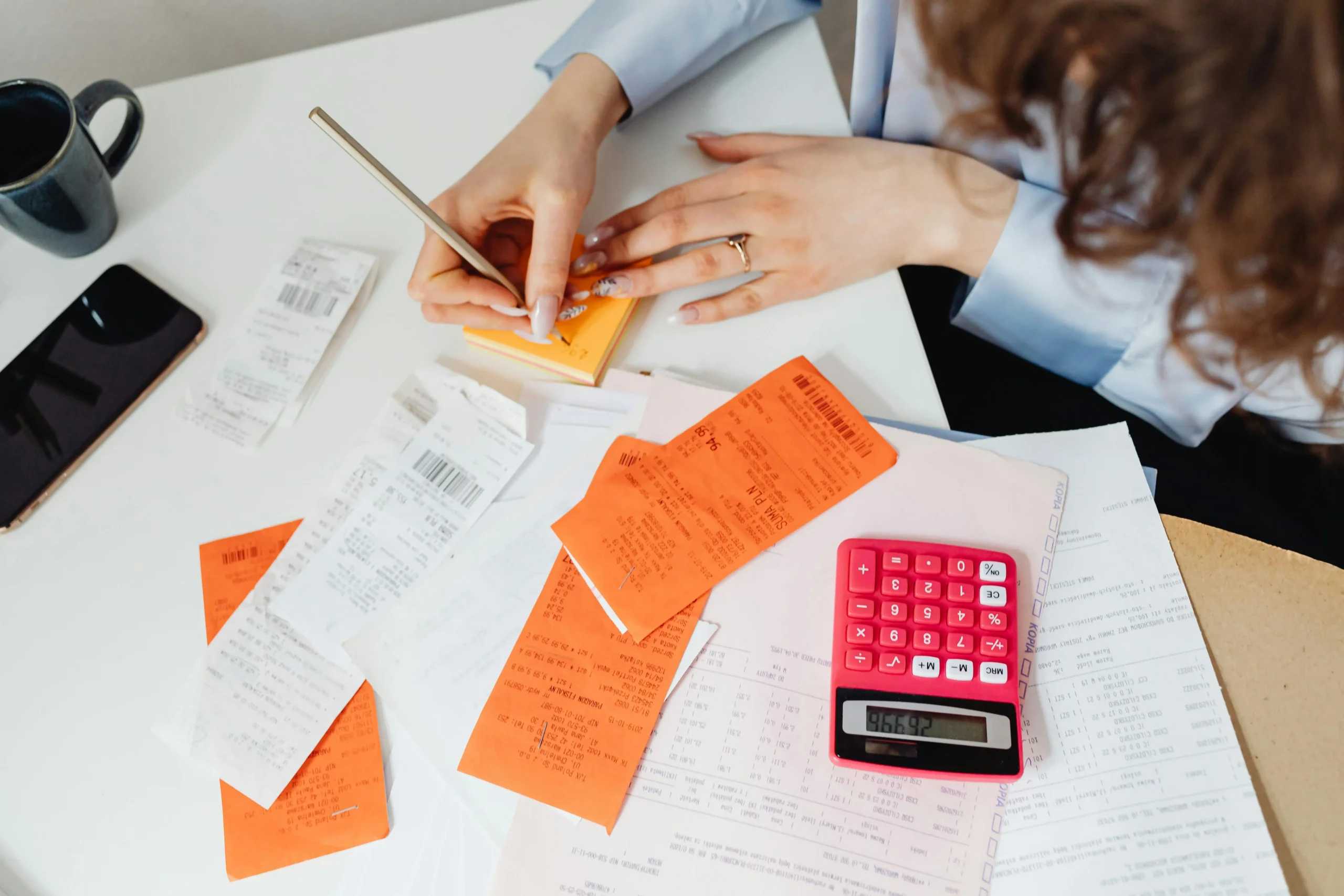 A person organizing receipts and invoices with a calculator, notebook, and smartphone on a desk, focused on managing finances.