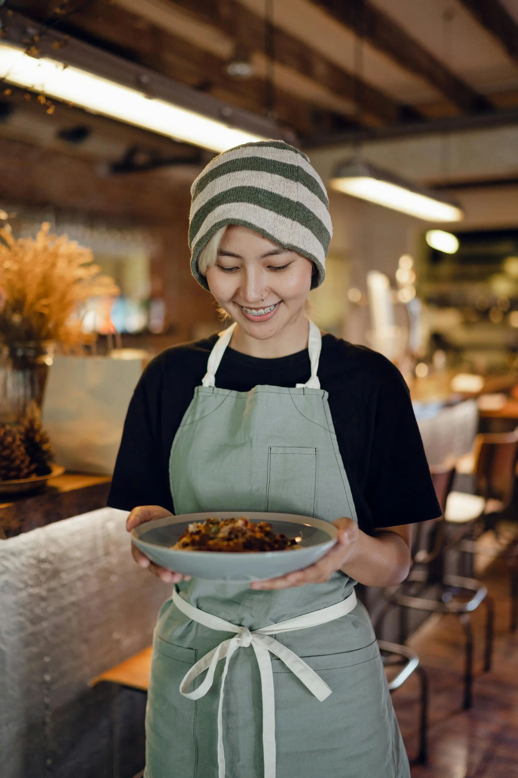 Woman wearing a striped headband and apron, smiling while holding a bowl of food in a warm kitchen setting