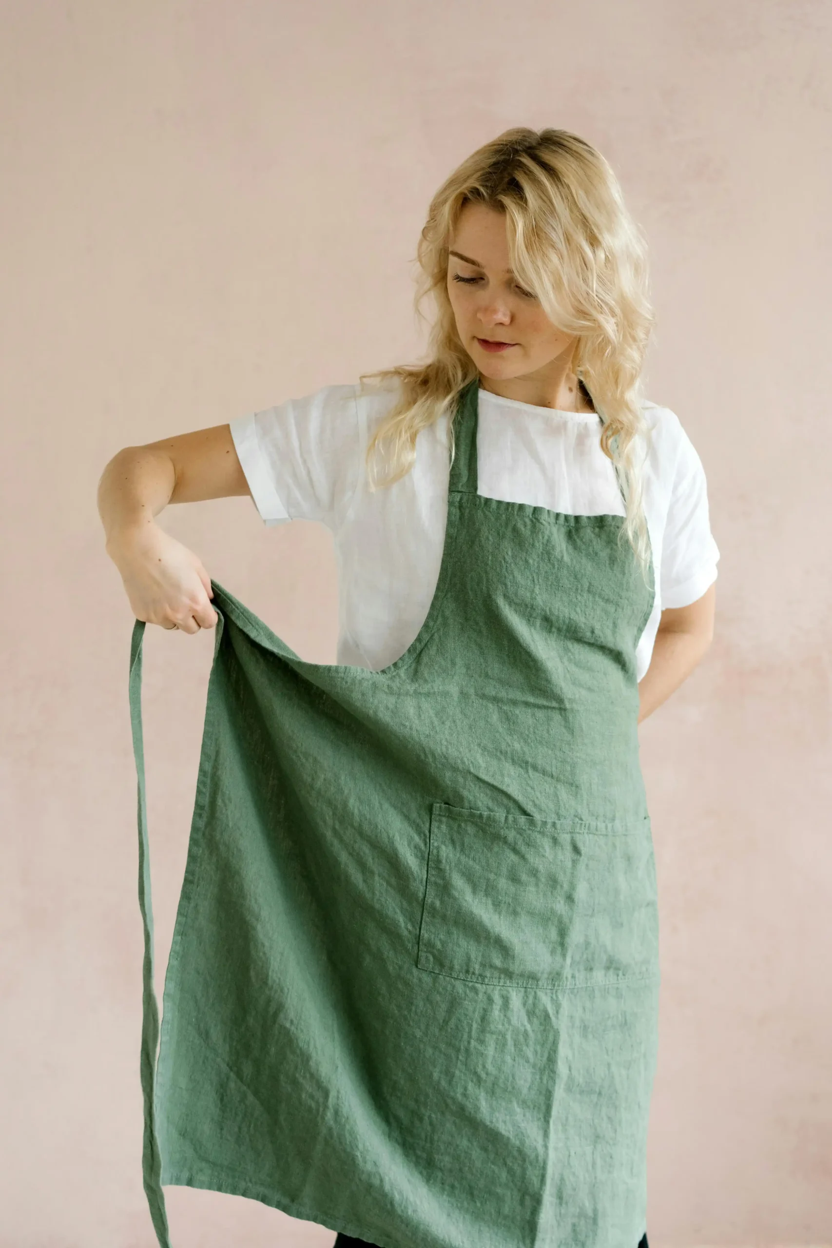 Woman tying a green apron while standing against a pink background, ready for cooking or baking.