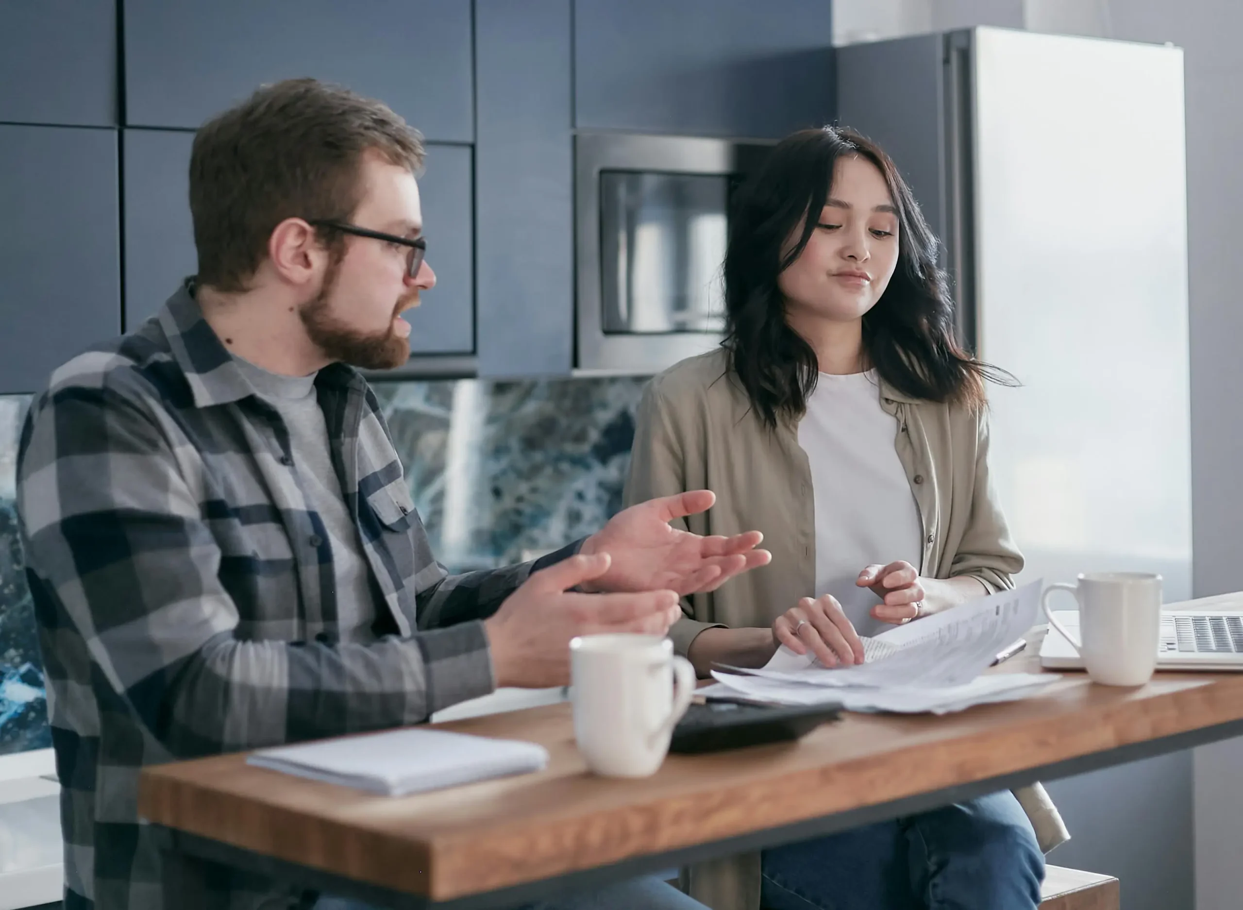 A man and a woman having a discussion over documents at a kitchen table with coffee mugs, laptops, and notebooks.
