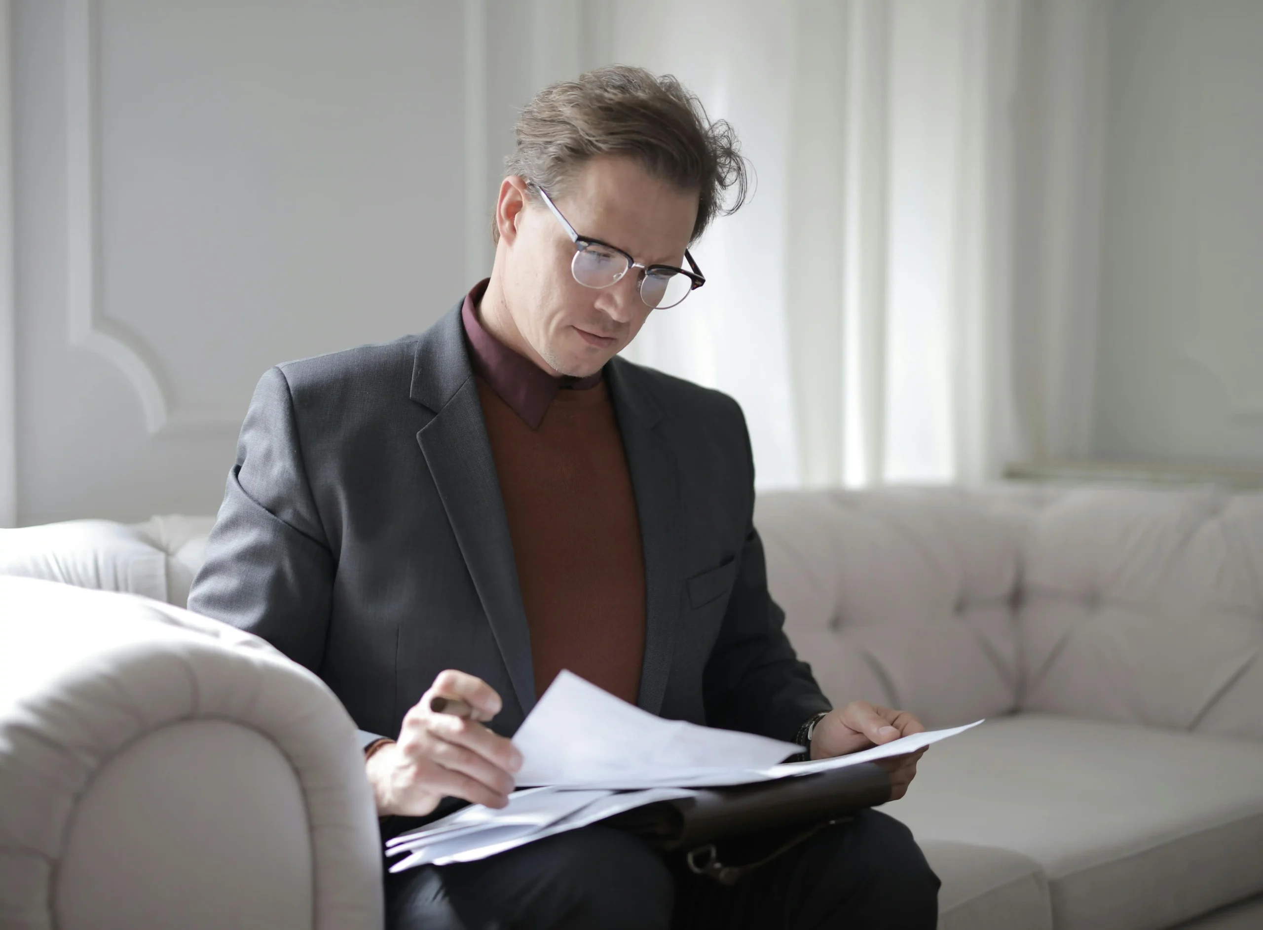 A man in a suit sitting on a sofa, reviewing papers with a focused expression.