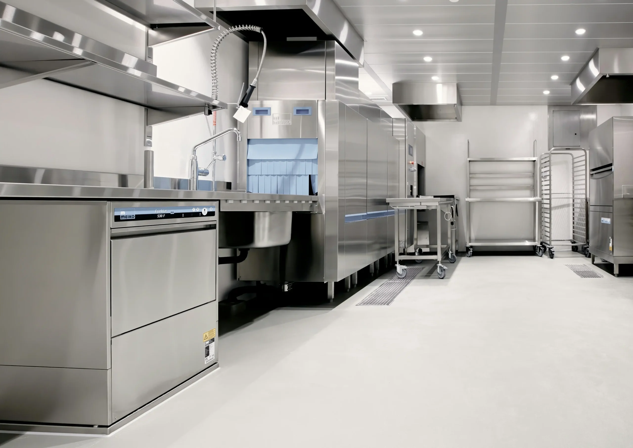 A professional, stainless steel commercial kitchen with a dishwasher, sink, oven, and storage racks.