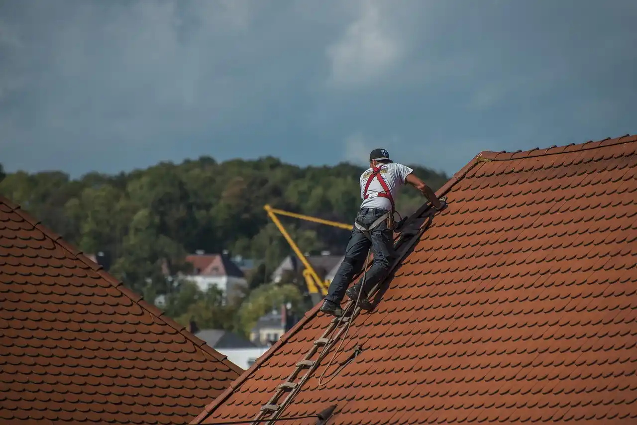 A roofer wearing safety harness and working on a steep roof using a ladder, with a crane and buildings in the background.